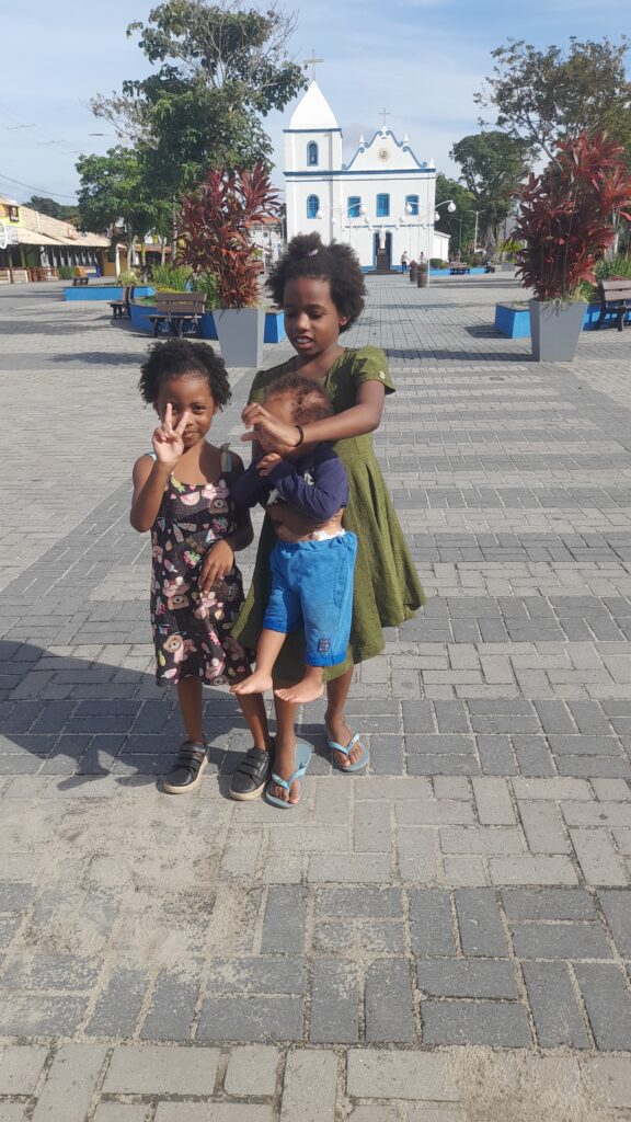 Three children stand on a paved plaza with a white and blue church in the background; a young girl in a floral dress makes a peace sign, while an older girl in a green dress holds a baby.