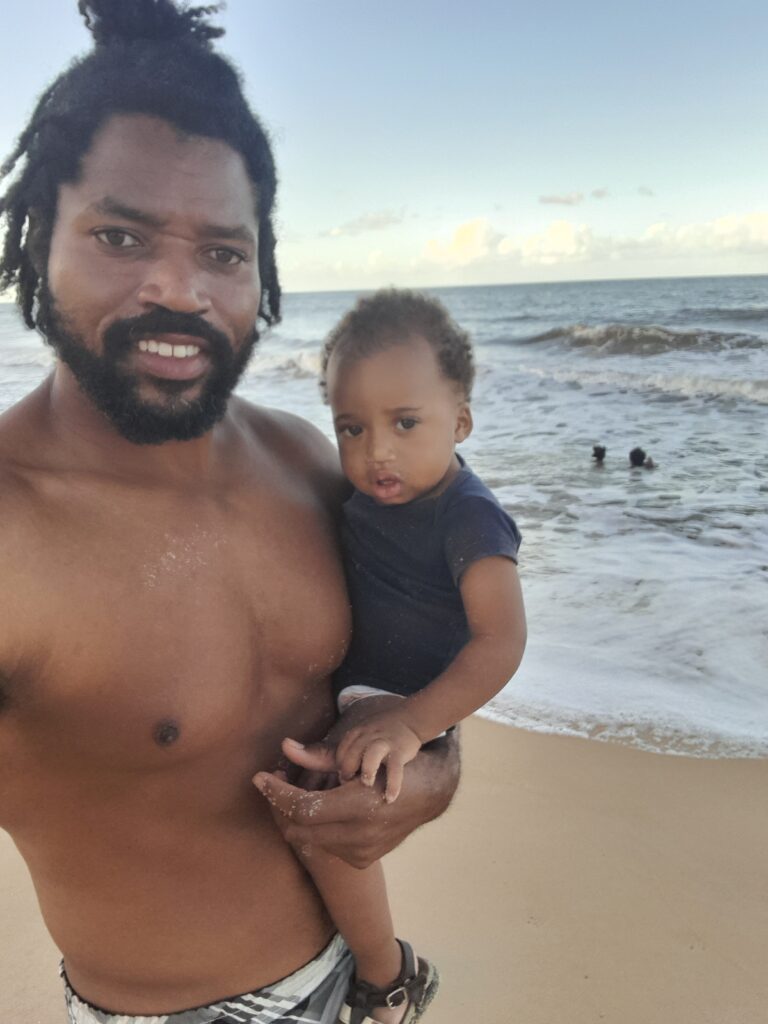 A smiling Black man with dreadlocks holds a baby on a sandy beach with ocean waves and a clear sky in the background.