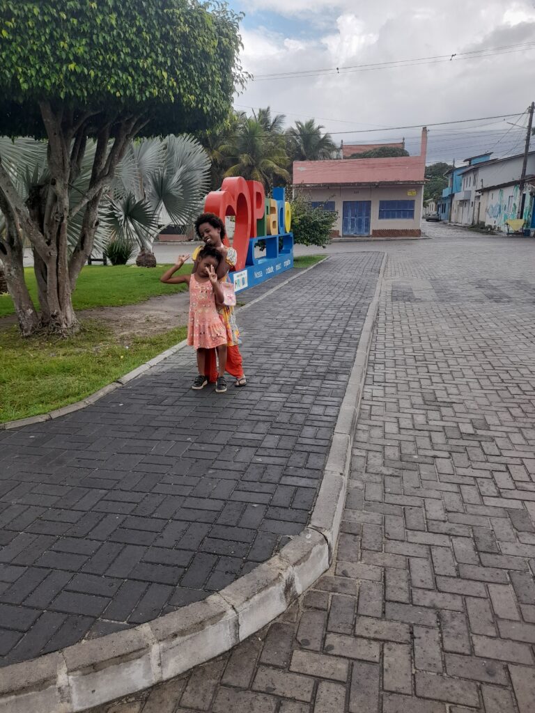 Two young girls pose for a photo on a paved walkway next to a large, colorful "COPAO" sign and a grassy area with trees, with a street and buildings in the background under a cloudy sky.