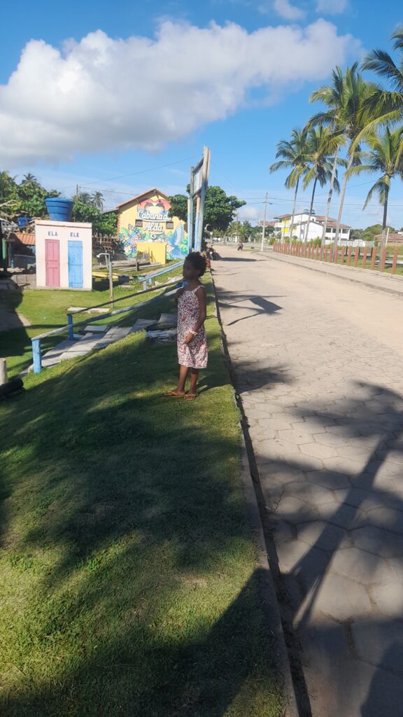 A young girl in a floral dress stands on a grassy area next to a paved road, with palm trees and colorful buildings, including one labeled "Cabanada Alda," under a bright blue sky.