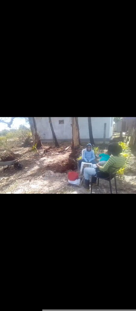 Two people sit on chairs, one in a light blue hoodie and the other in a striped shirt, facing each other outdoors with a red bucket between them