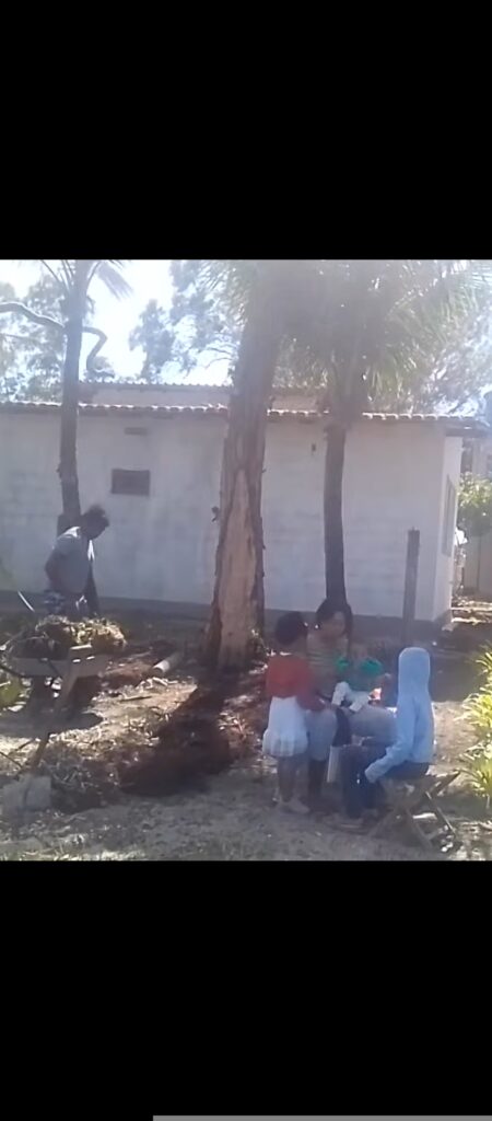 A man loads a wheelbarrow with dirt and debris while four children sit nearby, with a white building and palm trees in the background