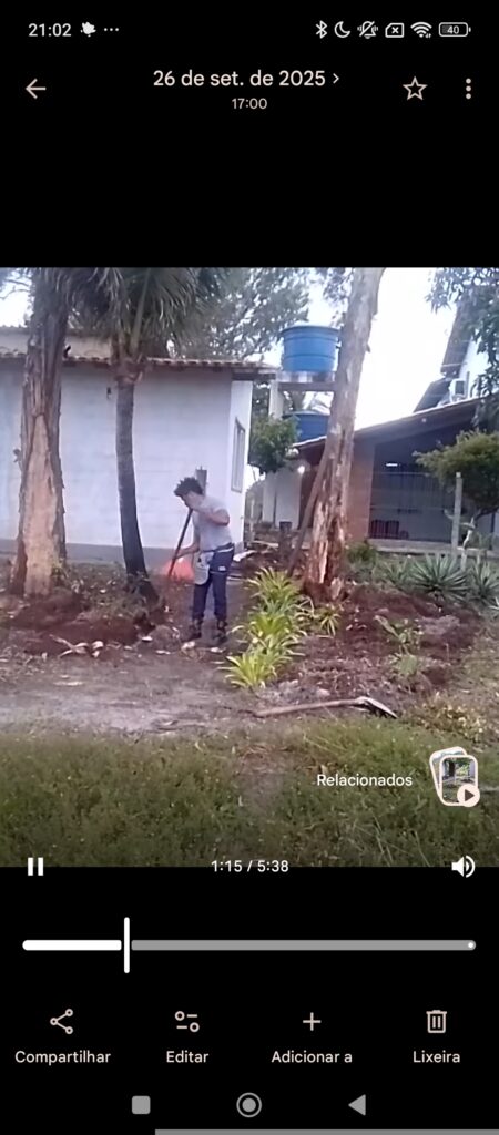 A person in a gray shirt and dark pants sweeps dirt with a broom in a yard with palm trees and a white building with a blue water tank