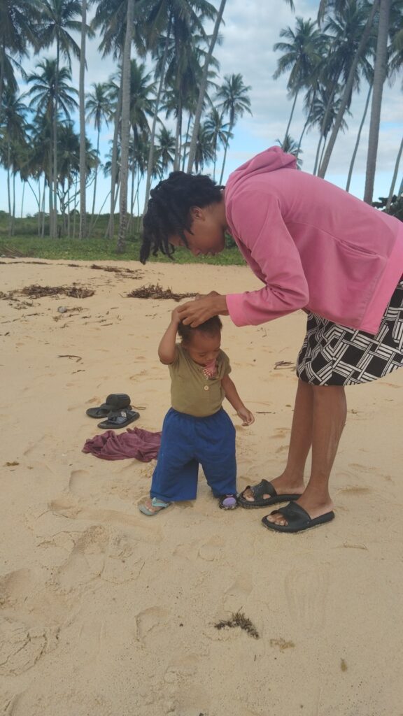 A person with dreadlocks in a pink hoodie bends over to help a toddler in blue pants stand on a sandy beach with palm trees in the background