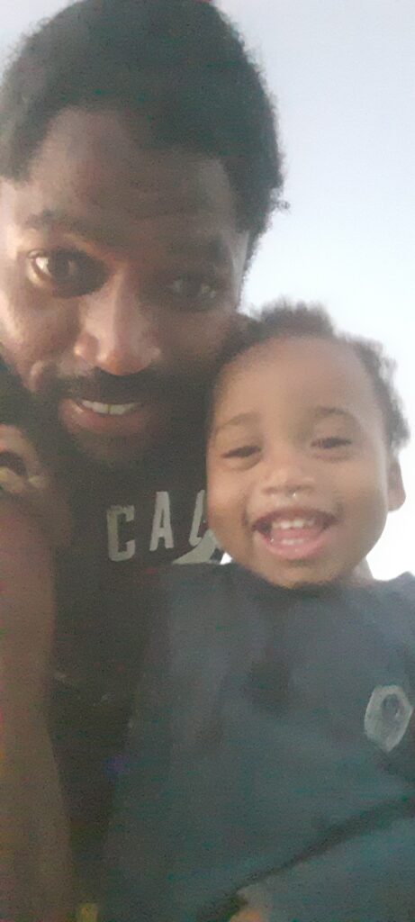 A smiling Black man with a beard and a black "CAL" shirt holds a happy young child with a nose ring, both looking directly at the camera