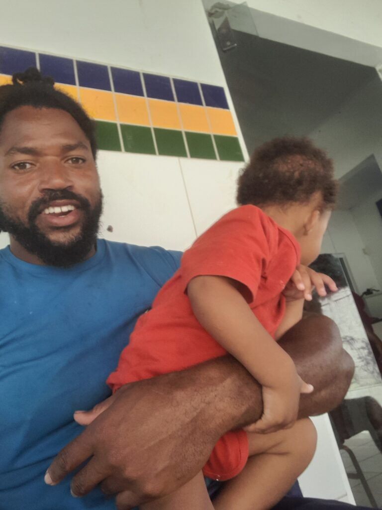A smiling Black man with dreadlocks and a beard holds a toddler in a red shirt, with a tiled wall featuring blue, yellow, and green stripes in the background