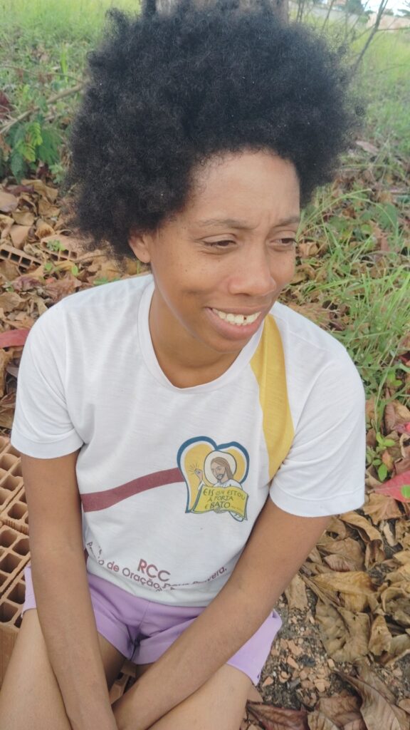 A smiling woman with dark, curly hair sits outdoors on a bed of dry leaves and dirt, wearing a white t-shirt with a religious graphic and light purple shorts