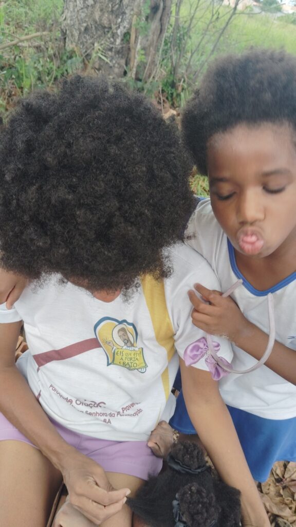 Two children with dark curly hair are outdoors; one child in a white shirt with a religious graphic puckers their lips, while the other child in a white shirt with "EIS QUE ESTÁ A PORTA E BATO" written on it looks down