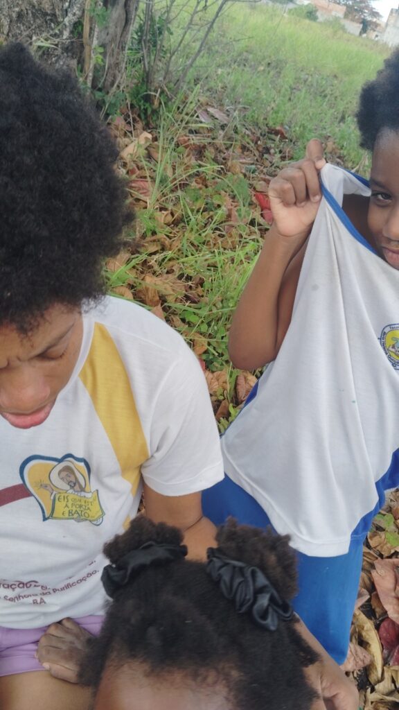 Two children are outdoors, one wearing a white shirt with a religious graphic that reads "EIS QUE ESTOU A PORTA E BATO," while another child in a blue and white shirt peeks from behind, with dry leaves and green grass in the background