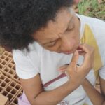 A person with dark curly hair and a white t-shirt with a yellow and red stripe sits next to a stack of terracotta bricks, looking down with a pensive expression