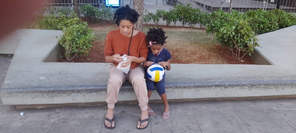A woman in an orange shirt and a young girl in a blue shirt sit on a concrete ledge, with the woman holding a white cloth and the girl holding a blue, white, and yellow volleyball