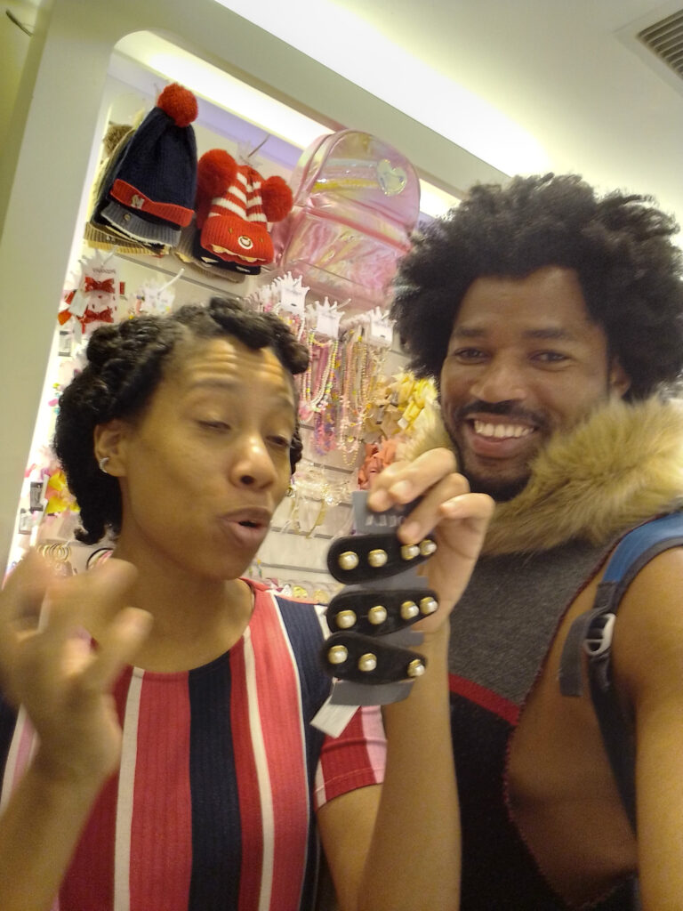 A smiling man with an afro holds up a black hair clip with pearls while a woman in a striped shirt looks on in a brightly lit store