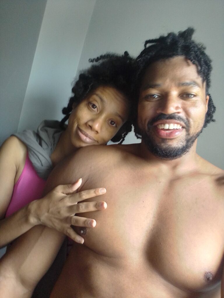 A smiling Black man with a beard and dreadlocks takes a selfie with a woman resting her head on his shoulder and her hand on his chest