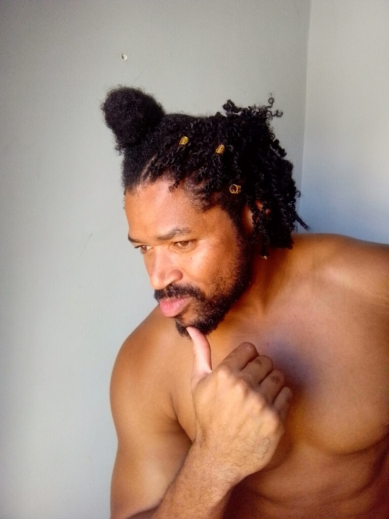 A shirtless man with a beard and dark curly hair, styled with a top bun and gold beads, looks down thoughtfully with his finger on his chin