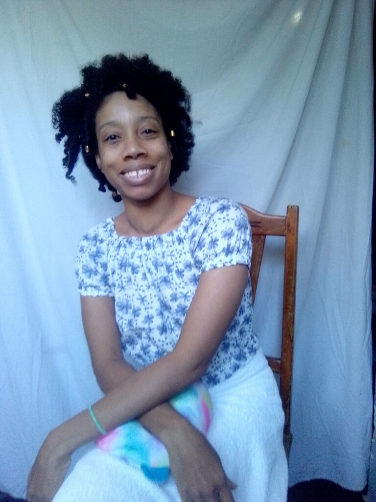 A smiling woman with dark, curly hair adorned with gold beads sits on a wooden chair, wearing a white and blue floral off-the-shoulder top and a white skirt, against a light blue draped fabric background