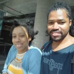 A smiling Black couple poses for a selfie under a concrete structure, with the man wearing a dark blue shirt with a light blue graphic that reads "76M e692