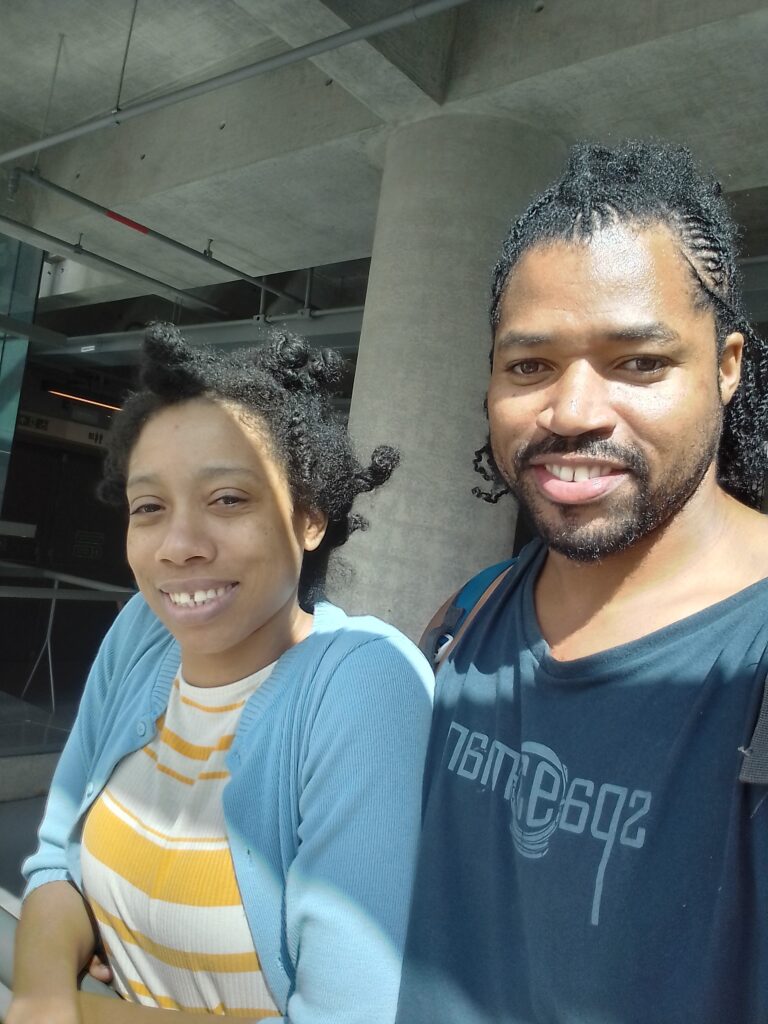 A smiling Black couple poses for a selfie under a concrete structure, with the man wearing a dark blue shirt with a light blue graphic that reads "76M e692