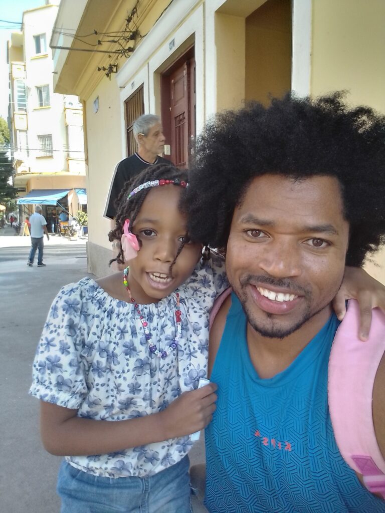A smiling man with an afro and a young girl with braided hair and a pink bow pose for a selfie on a city street