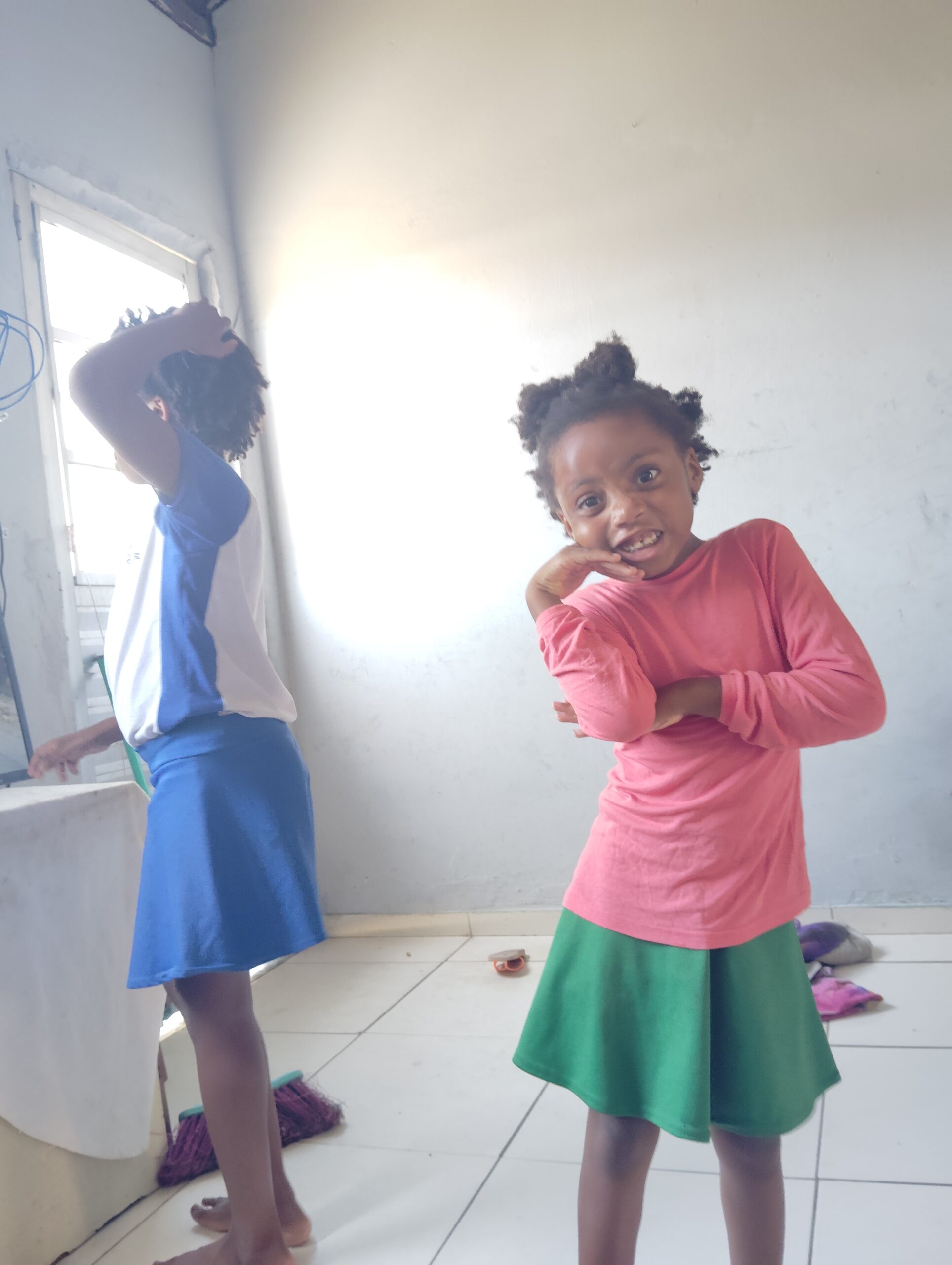 Two young girls with dark curly hair stand indoors; one in a pink shirt and green skirt smiles at the camera, while the other in a blue and white shirt and blue skirt looks out a bright window