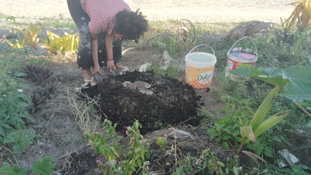 A woman with curly hair bends over, adding dark soil to a pile on the ground next to a white bucket labeled "Deline" and a red bucket