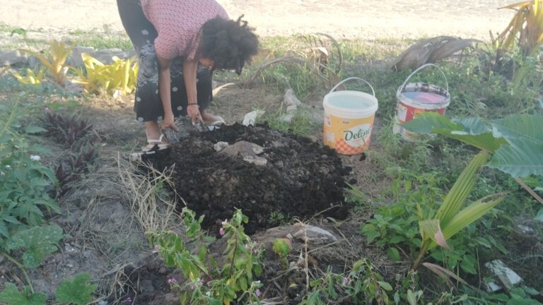 A woman with curly hair bends over, adding dark soil to a pile on the ground next to a white bucket labeled “Deline” and a red bucket