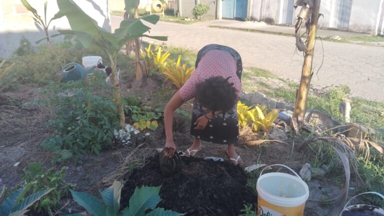 A woman with dark curly hair, wearing a red patterned shirt and black skirt, bends over to plant in a garden with dark soil, banana trees, and other plants