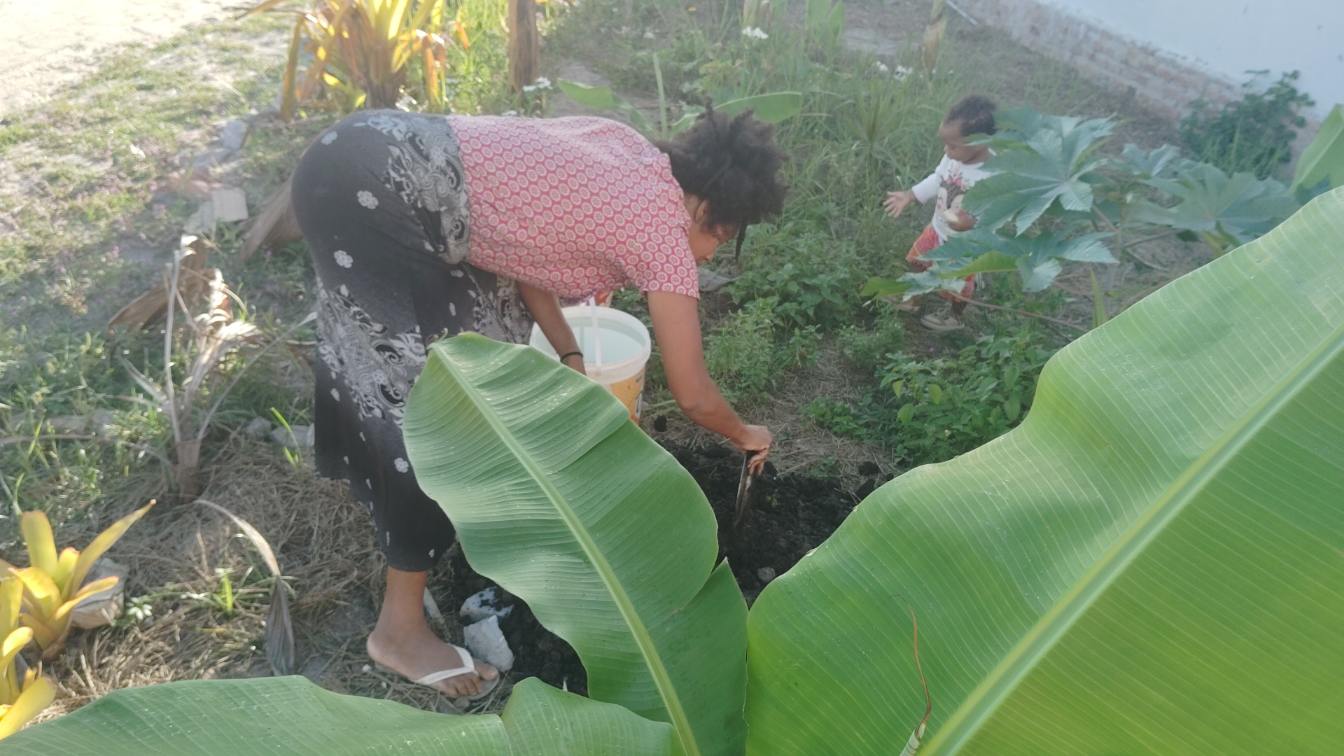 A woman with dark curly hair in a red patterned shirt and black skirt digs in a garden with a small shovel next to a white bucket, while a toddler in a white shirt plays in the background