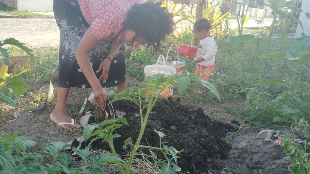 A woman in a patterned skirt and shirt kneels, digging in dark soil with her hands, while a toddler in patterned pants holds a red watering can nearby