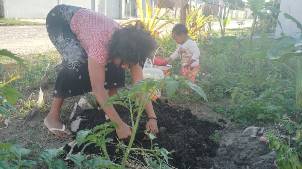 A woman with dark curly hair bends to plant a small tomato seedling in dark soil while a toddler in patterned pants waters plants with a red watering can in the background