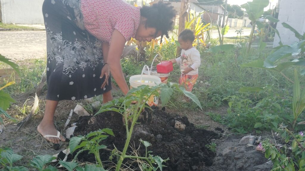 A woman in a patterned skirt and a toddler in colorful pants are gardening, with the woman bending over a mound of soil and the child holding a red watering can