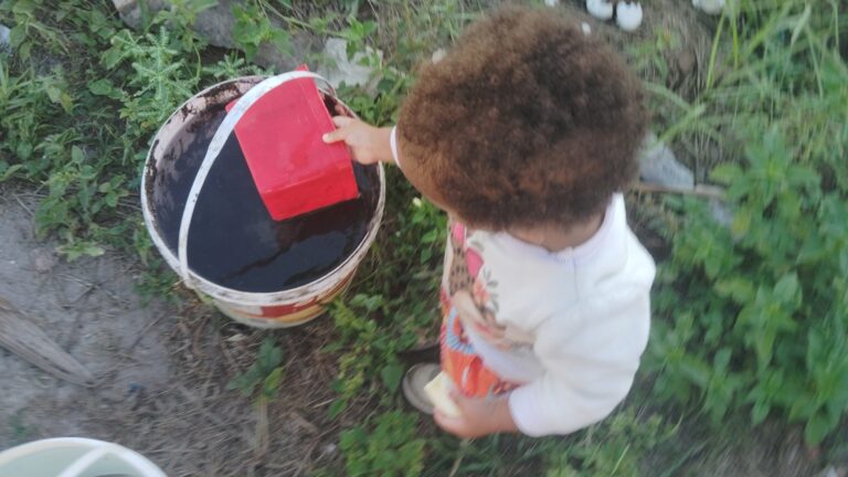 A young child with curly brown hair, wearing a white and patterned top, dips a red rectangular object into a bucket of dark water outdoors