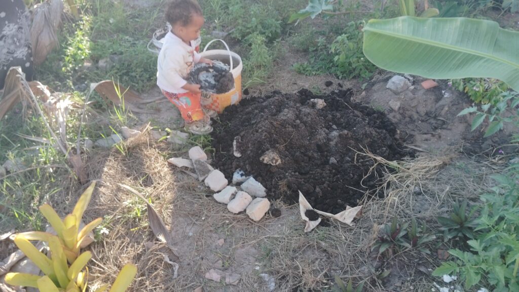 A small child in a white shirt and orange pants holds a bucket of dark soil, adding it to a large mound of dirt on the ground