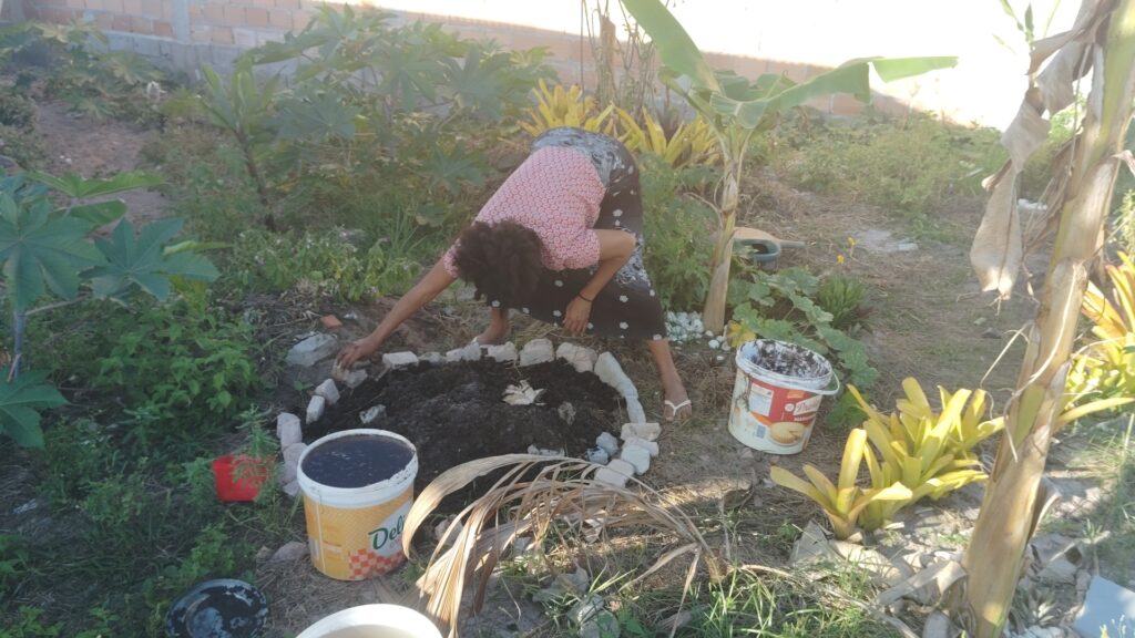 A woman with dark curly hair and a patterned shirt bends over a circular garden bed, arranging rocks around the edge of the dark soil