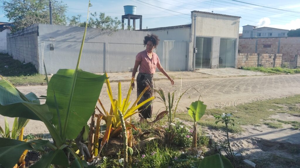 A woman with curly hair and sunglasses, wearing a patterned shirt and long skirt, stands on a paved street next to a small garden with banana plants and other foliage, with a light blue water tower visible in the background