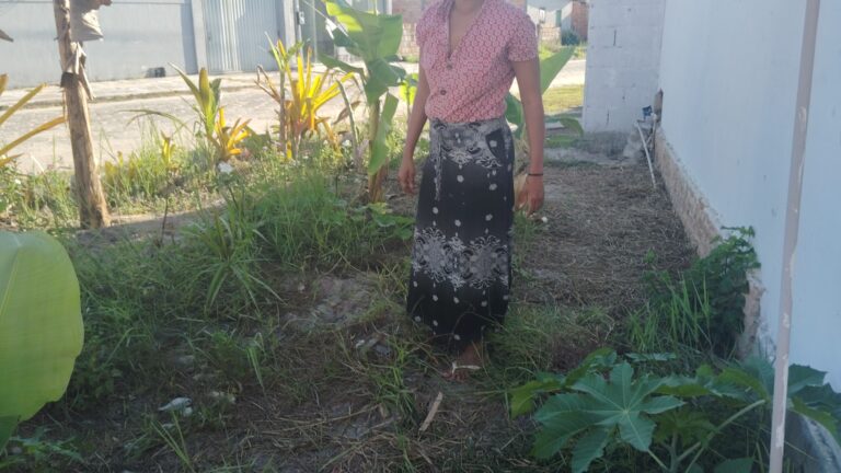 A person in a red patterned shirt and black patterned skirt stands in a grassy, overgrown yard with a white wall and banana plants