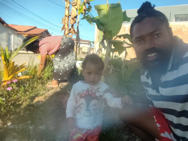 A man with a beard and dreadlocks smiles at the camera, holding a child in a white shirt with a deer graphic, while a woman tends to plants in the background