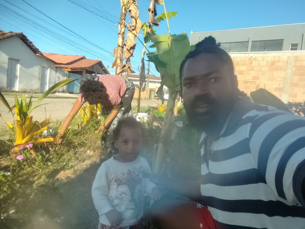 A man takes a selfie with a child and a woman gardening in a sunny, rural outdoor setting with houses and banana trees