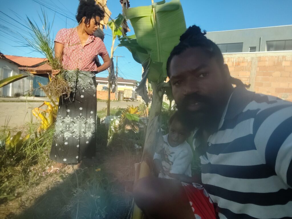 A woman in a patterned skirt holds a bundle of green plants while a man in a striped shirt takes a selfie with a child in a garden