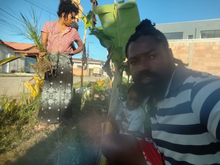 A woman in a patterned skirt holds a bundle of green plants while a man in a striped shirt takes a selfie with a child in a garden