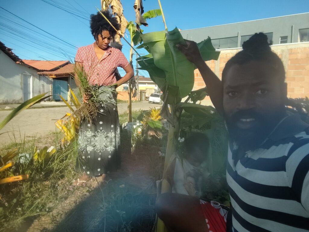 A man in a striped shirt takes a selfie while a woman in a patterned skirt and red shirt pulls weeds from the ground in a sunny, rural setting