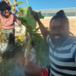 A smiling man in a striped shirt takes a selfie while a woman in a patterned skirt and shirt tends to plants in a sunny outdoor setting