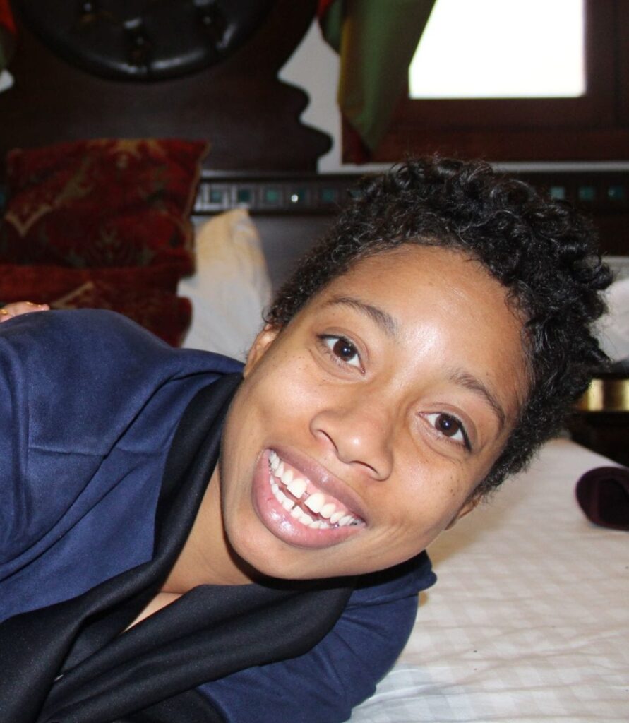 Sherika Trott Bailey, a woman with short curly hair, smiles broadly while lying on a white bed with a dark headboard in the background