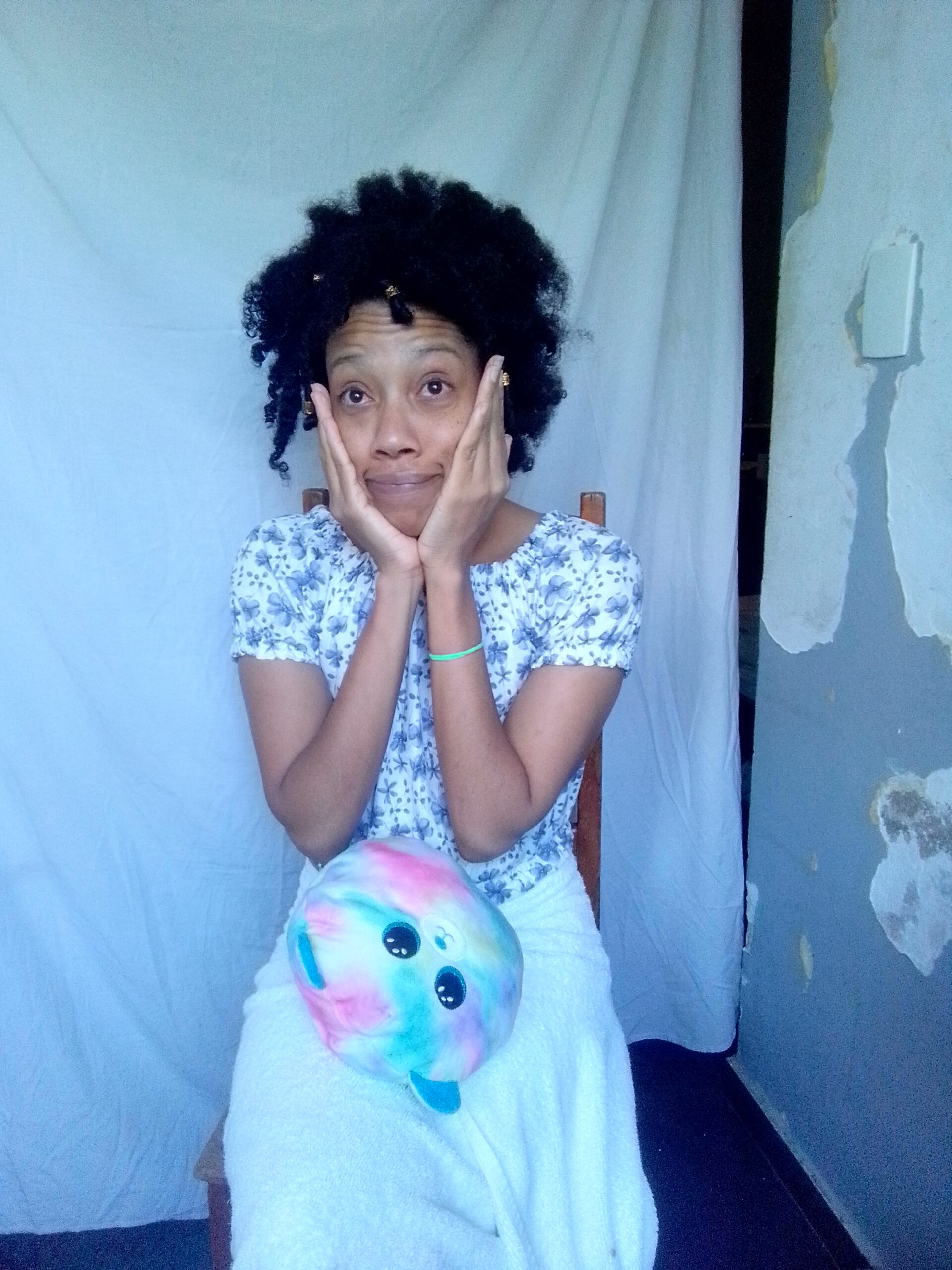 A young woman with curly dark hair and a floral top sits on a wooden chair, holding her hands to her cheeks with a surprised expression, a colorful plush toy resting on her lap.