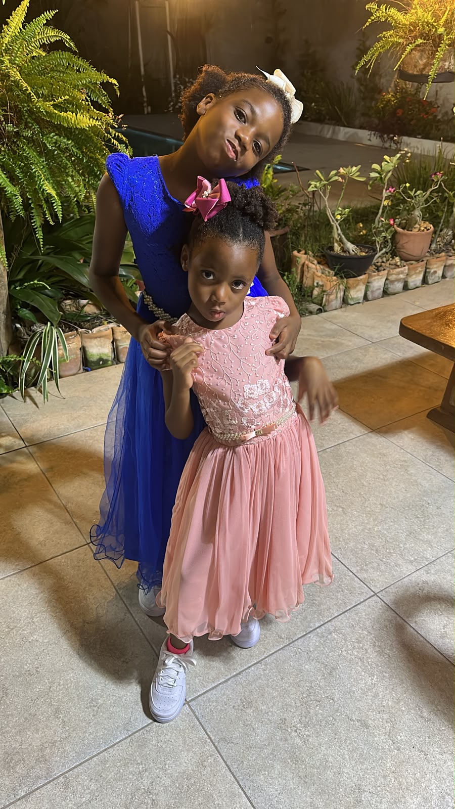 Two young girls, one in a blue dress and the other in a pink dress, stand together on a tiled patio at night, with potted plants in the background