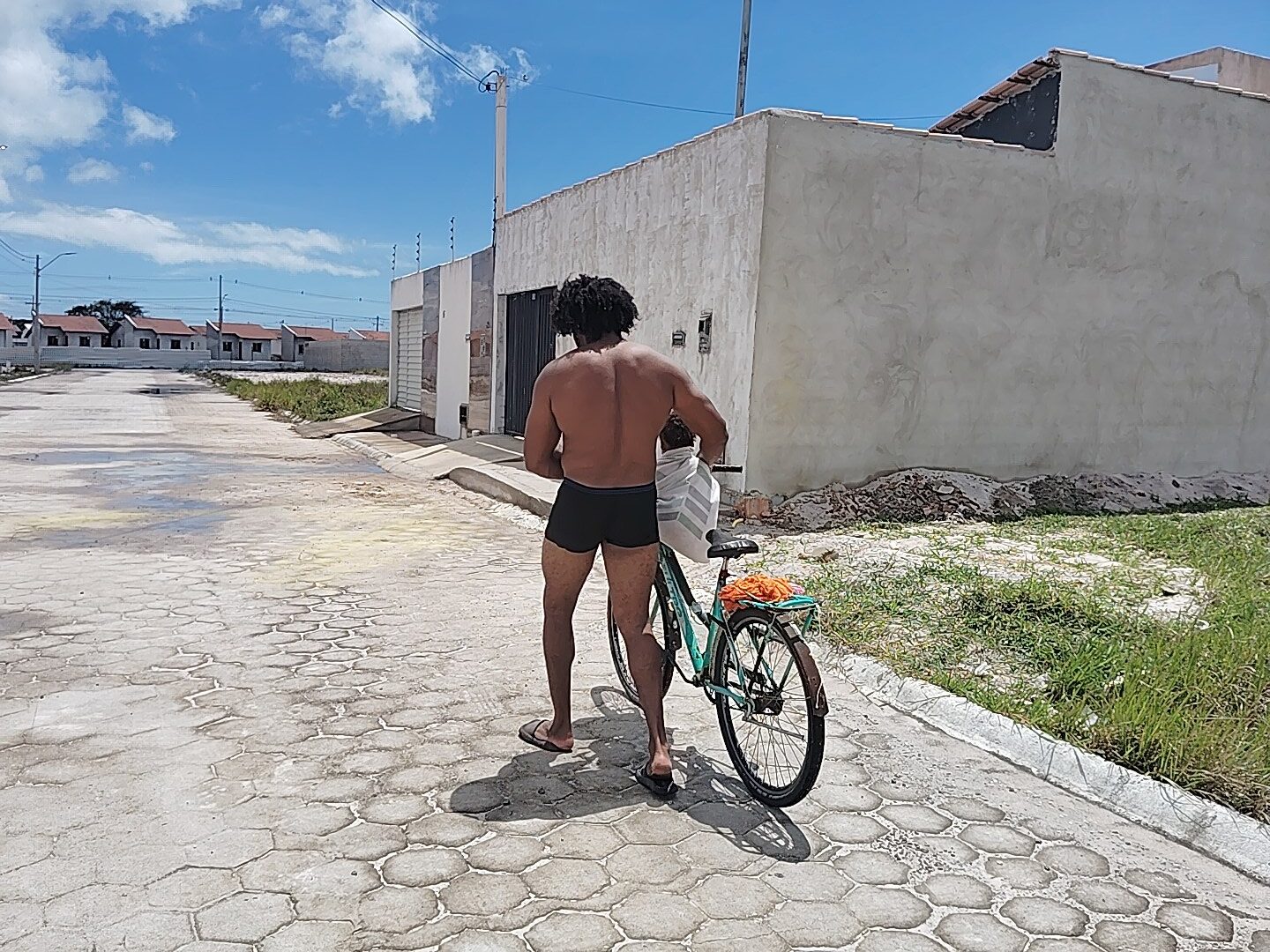 A shirtless man in black shorts and flip-flops pushes a teal bicycle with a white bag on the handlebars down a paved road